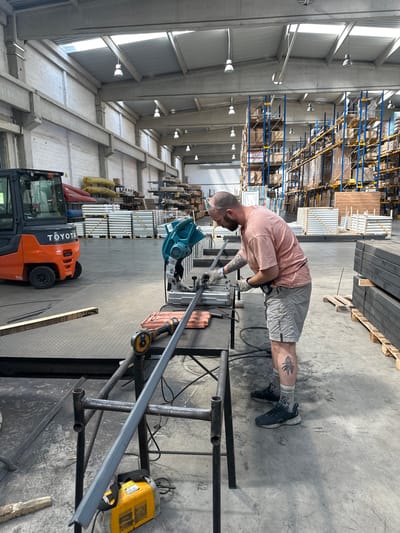 A man is using a chop saw to cut metal in a warehouse. The man is wearing a pink t-shirt, grey shorts, gloves, and safety glasses. He has tattoos on his arms and legs. In the background, there are shelves stacked with materials, a forklift, and a welding machine. The warehouse appears to be well-lit with natural light coming from the ceiling. The floor is concrete. The scene suggests a work in progress, with the man focused on his task. The location is Dub, Montenegro. The time of day is not explicitly stated, but the lighting suggests it is daytime.
