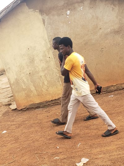 Two young men are walking side-by-side in Kachia, Nigeria. The man in the foreground is wearing a yellow t-shirt with an orange and white graphic design and light-colored pants. He is holding a black mobile phone in his right hand. The man behind him is wearing a dark t-shirt and brown pants. They are walking on a dirt path next to a light brown, textured wall. The environment appears to be outdoors with some debris on the ground. The wall has a rough, stucco-like finish with some discoloration and peeling paint in places. The ground is predominantly red-brown dirt with scattered dry grass and small white pieces of litter.