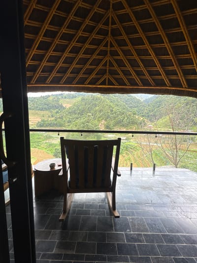 This image captures a serene outdoor balcony scene in Mù Cang Chải, Vietnam, offering a panoramic view of lush, terraced mountains. The vantage point is from inside a structure, looking out through a dark doorway onto a covered patio. Above, a striking, light-brown bamboo-framed roof with a thatched or woven material ceiling forms a precise grid pattern. The patio floor is paved with dark gray, rectangular stone tiles. A wooden rocking chair with blue cushions is positioned on the patio, facing away from the camera and towards the expansive landscape. Adjacent to the chair is a small, round wooden side table holding a light-colored bowl and a small red object. A clear glass railing with dark support posts provides an unobstructed view of the exterior. Beyond the railing, the landscape unfolds with numerous verdant, forested mountains and hills. Distinctive rice terraces, some appearing flooded and reflective, others showing dry, patterned earth, are carved into the mountain slopes. In the far distance, a valley floor is visible under an overcast sky, casting a soft, diffused light across the scene. A few bare-branched trees stand on the right side of the view. No people are visible.