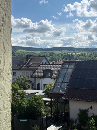 The image captures a bright, sunny daytime view of a residential area set against a backdrop of natural landscapes in Thüngersheim, Germany. The foreground features a blurred, textured concrete or stone wall, suggesting the photo was taken from an elevated position.
In the middle ground, several houses are visible, featuring dark tiled roofs. At least two of these houses are equipped with solar panels. A prominent architectural detail is a large, multi-story glass conservatory or sunroom attached to one of the houses, seamlessly integrated with its roofline. Lush green trees and bushes are abundant around the houses, and some potted plants can be seen near the conservatory.
The background unfolds into a wide expanse of green. Rolling hills covered with fields and meadows stretch towards a distant, dense line of dark green forest, marking the horizon. The sky is a vibrant blue, filled with large, fluffy white cumulus clouds, indicating clear and pleasant weather. On a black object in the lower right, the brand name "SONGMICS" is visible in light blue text. No people or specific activities are depicted in the scene.