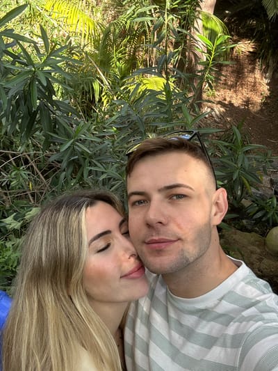 A man and a woman are lying together in what appears to be a garden or wooded area. The man has short brown hair and blue eyes, and he is wearing a striped t-shirt. The woman has long blonde hair and is lying with her eyes closed. They are surrounded by green foliage, including plants with long, narrow leaves and a palm-like tree in the background. It is difficult to determine the exact location of the photo in Altea, Spain without further information, but based on the vegetation, it is likely a botanical garden or park in the area.
