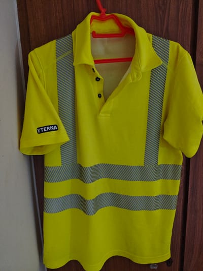 A bright yellow short-sleeved polo shirt with reflective silver stripes is hanging on a red plastic hanger. The shirt has a collar and a three-button placket. A black logo with white lettering that reads "TERNA" is visible on the left sleeve. The shirt is hanging against a background of a light-colored wall on the left and a dark wooden door on the right. The image appears to have been taken indoors, likely in a residential or storage setting. There is no discernible connection to the city of Athens, Greece.