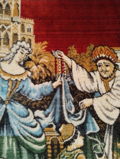 This image is a close-up photograph of a woven textile, identified as being located in Sucina, Spain. The textile artwork depicts a stylized scene featuring three human figures and architectural elements against a deep red background.
On the left side, a female figure is shown in profile, facing towards the right. She possesses light-colored, possibly white or pale blonde, hair styled upwards and wears a light blue gown with prominent folds and a decorative collar. Behind and above her, a tan-colored building with multiple arched windows and turrets, resembling a castle or large manor, is visible.
On the right side, a male figure faces towards the left. He has a beard and is adorned with an orange and brown headpiece. He wears a white or light-colored robe with a gold trim around the neckline. His right arm is extended towards the center, holding a multi-strand beaded necklace composed of red and white elements.
The female figure's hands are positioned to receive this necklace from the male figure, indicating an exchange. Between these two primary figures, stylized brown and gold foliage or trees are depicted. At the bottom center of the frame, a partial view of a smaller, possibly kneeling figure, dressed in blue and green attire, is observable. The upper portion of the background consists of a solid deep red color. The texture of the image is characteristic of a coarse, woven fabric.