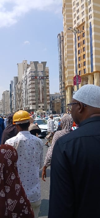 The image depicts a bustling street scene in Makkah Al Mukarramah, Saudi Arabia, under a clear, bright sky. In the foreground, viewed from behind, a male figure on the right wears a white knitted kufi and dark-rimmed glasses over a black shirt. To his left, another male figure wears a bright yellow hard hat over a dark head covering and a white long-sleeved shirt with a subtle dark pattern. A third individual is partially visible on the far left, wearing a maroon garment with a white repeating pattern.
The midground is densely populated with numerous pedestrians, including men and women dressed in various attire, navigating the street. Several white passenger vehicles are present on the thoroughfare, interspersed among the crowd.
The background features a dense urban environment dominated by multiple tall, multi-story buildings. On the right, a prominent building exhibits a light yellow and brown striped facade with vertical blue-tinted window panels and ornate architectural detailing. An elaborate street lamp with multiple arms is affixed to this building. To the left and extending into the distance, a succession of other high-rise buildings, predominantly in light colors such as white, grey, and beige, line the street, characterized by numerous windows. The sky above is clear blue with minimal scattered white clouds. The scene conveys active pedestrian and vehicular movement within a developed urban setting.