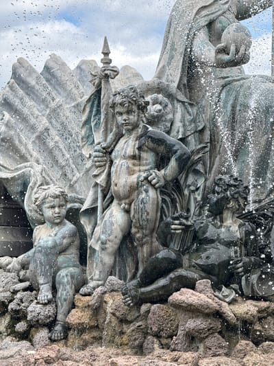 This is a close-up, eye-level shot of a bronze fountain in Bordeaux, France, depicting several sculpted figures, possibly cherubs or putti, interacting with water. The figures appear weathered, with a patina indicating age. The central figure is a boy standing, holding a staff. To his left, a smaller child sits. To the right, another child is depicted reclining, holding an object. Water is actively spraying and splashing around the sculptures, creating a dynamic and wet environment. The background shows stylized, rocky formations and a cloudy sky. The fountain is situated amidst a rocky, uneven base.