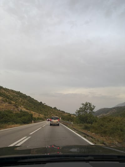 The image depicts a two-lane road in the city of Cerovo, Montenegro, on an overcast day. The perspective is from inside a vehicle, looking forward. The road surface appears wet, indicating recent rain. Two cars are visible on the road ahead: a silver SUV in the right lane and a red car further ahead in the left lane. The road is bordered by a steep, green, and rocky hillside to the left and a dense line of trees and bushes to the right. The sky is uniformly gray and cloudy. The foreground shows the hood and windshield of the vehicle from which the photo is taken.