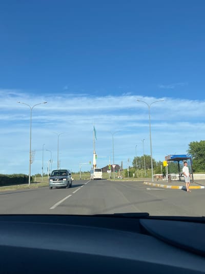 The image depicts a street scene in Stepnogorsk, Kazakhstan, likely during the daytime given the bright sunlight. The perspective suggests the photo was taken from inside a vehicle, looking out.
The foreground is dominated by the dark interior of the vehicle, likely a dashboard or door panel. Beyond this, a paved road stretches into the distance, lined with several streetlights and some greenery. The sky is a clear, bright blue with wispy white clouds.
On the right side of the road, a bus stop shelter is visible. A person, wearing a white t-shirt and light-colored shorts, is standing at the bus stop. Their body language doesn't convey a strong emotion. There is some text on the blue sign of the bus stop, but it is not legible in the image. Yellow and white striped barriers are present near the bus stop.
Several vehicles are visible on the road. A dark-colored van is driving away from the camera in the middle distance. Further ahead, a white vehicle, possibly a bus, is also on the road. Traffic lights are visible, showing green. The presence of the bus stop and the road suggests an urban or suburban setting within Stepnogorsk. The overall atmosphere appears to be calm and ordinary, with no indication of any particular event or urgency.