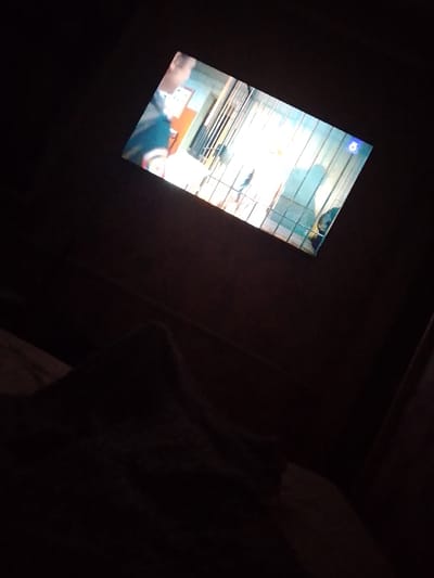 The image shows a television screen displaying a scene with people and some objects. The screen is angled, and the surrounding environment is dark, suggesting the photo was taken in a dimly lit room at night. On the screen, there appears to be a person in the foreground on the left, wearing a dark top with colorful stripes on the sleeves. The background on the screen shows a metal grate or bars, possibly indicating a jail or a similar enclosed space. There's a bright light source behind the grate, and some indistinct figures are visible beyond it. In the bottom right corner of the TV screen, there is a blue logo with a white outline of a figure, possibly a channel logo or a graphic element. The foreground of the image is very dark, with the indistinct shape of a blanket or bedding visible at the bottom. The overall impression is of someone watching television in a dark room.