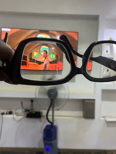 The image prominently features a pair of black-framed eyeglasses held in the foreground, slightly angled. Through the lower lens of the eyeglasses, a clear reflection of a person's face, likely male, is visible, surrounded by what appears to be a cockpit or control panel with circular green and orange displays, and a hand. A similar, but out-of-focus, rectangular screen or poster displaying the same cockpit scene with an orange hue is visible in the background to the left, outside the glasses.

The background to the right of the eyeglasses reveals a light-colored wall. Mounted on or near this wall is a small, grey-bladed fan, which shows motion blur, indicating it is running. Below and to the right of the fan, various electronic devices are present, including a blue unit with a prominent glowing blue indicator light and a smaller black box with a green light, connected by wires. The setting is indoors, likely a room, with artificial lighting. No discernible text is visible on any objects or reflections within the image. The context provided is Nev, Nigeria.