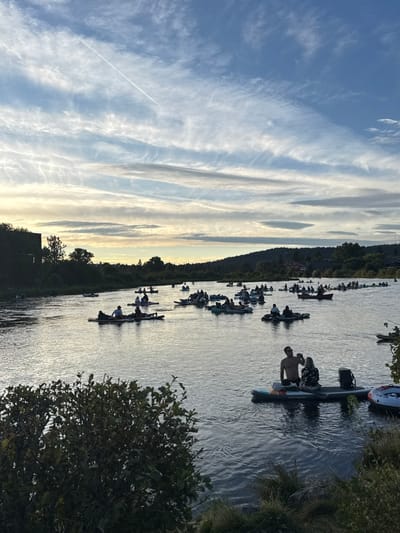 This image depicts a wide-angle view of the Deschutes River in Bend, Oregon, United States, during late afternoon or early evening. The river is densely populated with approximately 70 to 100 individuals engaged in recreational water activities.
**People:** Participants are distributed across numerous personal flotation devices, predominantly inflatable stand-up paddleboards and various inflatable rafts or tubes. They are observed in groups ranging from one to four people per vessel, largely in a relaxed, seated, or reclining posture, indicating a leisurely float. In the foreground right, two individuals are prominent on a large inflatable paddleboard: a shirtless male facing away and a female in a patterned top seated facing left, appearing to interact.
**Objects:** The watercraft consist mainly of inflatable paddleboards and rafts, with some appearing to be inflatable rings or tubes. A dark cooler is visible behind the two individuals in the foreground. Paddles are discernible on some vessels.
**Actions and Interactions:** The primary action is recreational floating down the river. Interactions between individuals on the same or adjacent vessels are implied by proximity and body orientation.
**Environment:** The river's surface is relatively calm, displaying subtle ripples and reflections of the sky and light. The riverbanks are heavily vegetated, with dense green shrubs in the immediate foreground on the left. Distant riverbanks are characterized by forested hillsides, with some indeterminate structures visible amongst the trees on the far right. A dark, rectangular structure, possibly part of a bridge or building, is partially visible on the far left bank.
**Sky:** The sky features a clear blue expanse with extensive, horizontally elongated, wispy cirrus and cirrocumulus clouds. A distinct, linear white contrail extends from the upper left quadrant towards the mid-right. The horizon line exhibits a soft, luminous band of lighter, golden-white light, consistent with low sun angles at dusk.