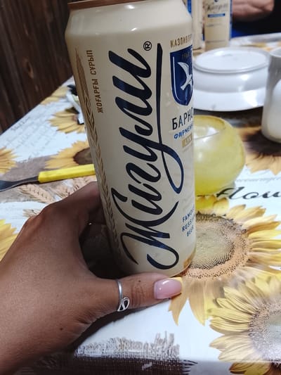 A hand with pink nail polish and a silver ring is holding a can of "Zhuzhaly" beer. The can is light beige with dark blue lettering. The label also features a blue crest with a white bird and text that reads "КАЗПИВПР", "1963", "BARNO", "ФИРМЕННОЕ", "4.9%", and "FAMOUS RUSSIAN BEER". The background is a table with a tablecloth featuring sunflowers. There are other items on the table, including a yellow utensil and what appears to be another beverage container. The lighting suggests it could be daytime, with a relaxed atmosphere.