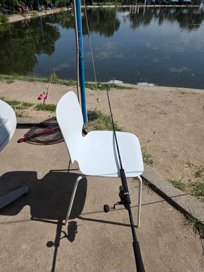 In German, Bulgaria, on a sunny day, a fishing setup is arranged by a lake. In the foreground, a white modern chair is positioned on a concrete ground. A fishing rod with a reel is placed on the chair. To the left of the chair, a white round table is visible, and a fishing net with a red handle lies on the ground next to it. Further back, a pink children's fishing rod is on the sandy bank. In the background, the lake reflects the surrounding trees and the sky. Several people are seated on benches along the opposite bank, some under umbrellas. The environment is a recreational fishing area.