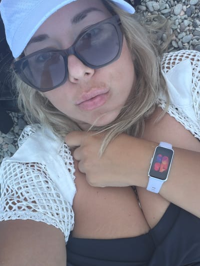 A woman lies on a bed of grey pebbles, her body angled towards the viewer. She wears a white mesh top over a dark swimsuit, and a white cap partially obscures her blonde hair. Large, dark sunglasses cover her eyes, and her lips are slightly parted. On her left wrist, she wears a light purple smartwatch displaying a colorful abstract graphic. The scene appears to be outdoors, given the pebbles and natural lighting, suggesting a beach or rocky shoreline. The overall impression is one of relaxation and leisure, possibly during daylight hours. There is no visible text in the image.