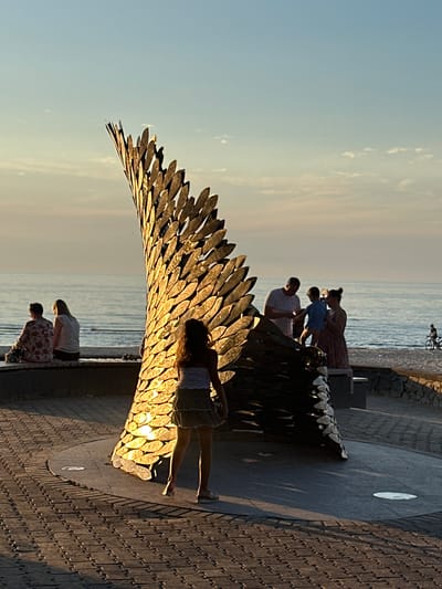 A family of four, including two adults and two children, are enjoying a sunset by the sea. The father, wearing a white t-shirt, is holding his son, who is wearing a blue shirt, while the mother, in a patterned dress, stands beside them, looking at them. They are standing on a paved promenade, with a large, metallic sculpture resembling a wing in the foreground. The sculpture is catching the warm, golden light of the setting sun, creating a beautiful visual effect. In the background, the sea stretches out to the horizon, with a few clouds in the sky. The atmosphere is peaceful and serene, with the family enjoying a quiet moment together. The image is taken in Jūrmala, Latvia, and the time of day is sunset. There is no visible text in the image.