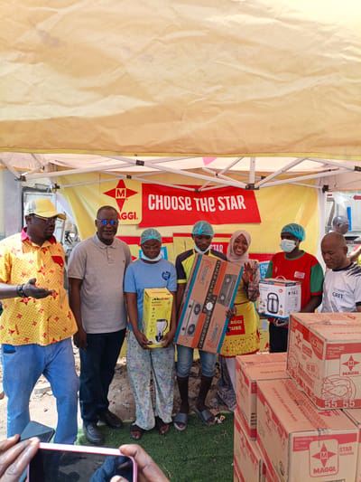 The image captures a promotional event, likely outdoors under a yellow tent, in Tarauni, Nigeria. A group of seven people, appearing to be a mix of staff and customers, are posing for a photo. The subjects are diverse in age and gender, with most wearing casual clothing. Some individuals are wearing hairnets, suggesting they might be involved in food preparation or distribution.

The central focus of the event seems to be on Maggi products, as evidenced by prominent Maggi branding on boxes and a banner. The banner reads "CHOOSE THE STAR" with the Maggi logo. Several cardboard boxes, stacked high, are visible in the foreground and background, displaying text like "MAGGI CHICKEN," "STAR SEASONING," and "STORE IN A COOL AND DRY PLACE."

The people are holding and displaying several products, including a blender and grinder (Mewe brand), a gas stove (Bossan brand), and an electric kettle (Boscon brand). One man on the far left is gesturing with his hand while speaking, possibly explaining the products or the promotion. The mood appears positive and celebratory, with smiles on some faces, particularly the woman in the center who is waving.

The ground is a mix of sand and green grass, and the lighting suggests it is daytime. The overall scene suggests a community-focused promotional activity or a distribution event for Maggi products in a public or semi-public outdoor space in Nigeria.