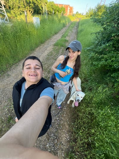 A selfie photograph shows two individuals and a white dog on a dirt road in Lozarevo, Bulgaria. The individual in the foreground is smiling and holding the camera. This person is wearing a black vest over a blue shirt. Behind this person is a woman wearing a blue top, grey pants, and a grey baseball cap. She is holding a small bouquet of wildflowers and has her hand on the white dog, who is panting. The dirt road stretches into the distance and is flanked by green grass and vegetation. Buildings are visible in the background.