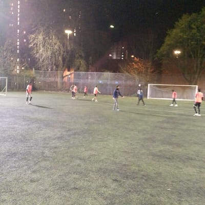An overhead, low-light photograph depicts an active nighttime football match or practice on an artificial turf pitch in Coventry, United Kingdom. Approximately ten individuals, primarily males, are distributed across the field, wearing various athletic attire including orange, white, blue, and dark colored tops. Two white football goals with nets are present at opposing ends of the pitch. A football is visible airborne near the right side of the frame. The field is illuminated by several overhead floodlights, casting shadows. In the background, beyond a chain-link fence, bare trees and illuminated windows of multi-story buildings are discernible, indicating an urban setting.