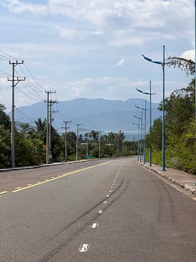 This is a vertical, eye-level, medium shot of a paved road in Vĩnh Lương, Vietnam, with a mountainous landscape in the background. The road has a yellow double line marking its center and white dashed lines marking the right edge. Tire marks are visible on the asphalt. On the left side of the road, there are multiple utility poles with wires. On the right side, there is a sidewalk with a row of distinctive blue lampposts. Lush green trees and vegetation line the right side of the road. The sky is partly cloudy. There are no people or animals visible in the image.