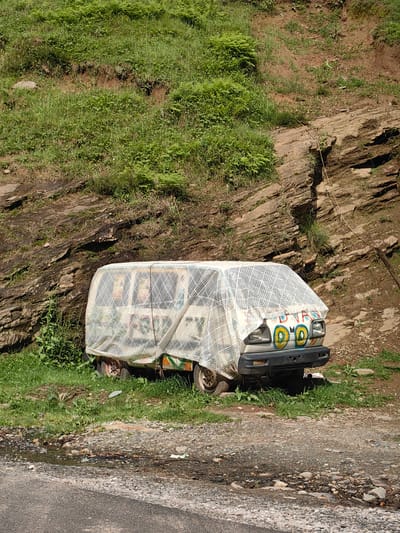 A white van is covered by a white patterned tarp and parked on the side of a dirt road. The van is decorated with colorful drawings on the front. The background consists of a steep, rocky hill covered in green grass and vegetation. There is a small puddle of water and some debris on the road in front of the van. The scene appears to be in Kothi, India.