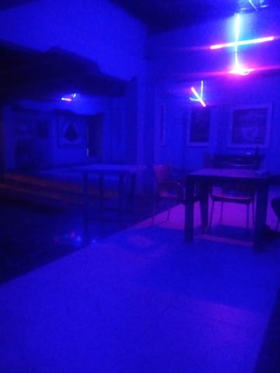 The image depicts an interior scene illuminated by blue and pink neon lights, creating a dark and atmospheric environment. In the foreground, a large, flat, dark-colored surface, possibly a floor or table, dominates the lower portion of the frame. Several dark-colored chairs are positioned around a dark-colored table in the middle ground, suggesting a seating area. In the background, more furniture and what appear to be framed pictures or displays are visible on a wall, all rendered in deep blue tones due to the lighting. Suspended from the ceiling are bright neon light fixtures, one forming a cross shape with pink and white elements, and another, smaller, V-shaped fixture. The overall impression is that of a dimly lit establishment, possibly a lounge, bar, or club, in Sabon Gari Nassarawa, Nigeria, where the pervasive neon lighting significantly influences the visibility and color of all objects. There are no people clearly visible in the image.