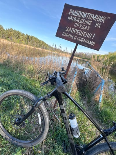 The image depicts a black Trek bicycle lying on its side in a grassy area next to a body of water, likely a pond or lake. A weathered, brown sign with white Cyrillic text stands nearby. Behind the sign and bicycle, reeds and vegetation line the water's edge. A blue, metal structure or barrier is partially visible behind the reeds. In the background, a treeline is visible against a blue sky with light clouds. The location is Капцёўскі сельскі Савет, Belarus, as indicated by the Cyrillic text. The sign likely conveys a message regarding the fish farm and restrictions or regulations related to the area, possibly prohibiting dogs. The bicycle appears to be unattended.