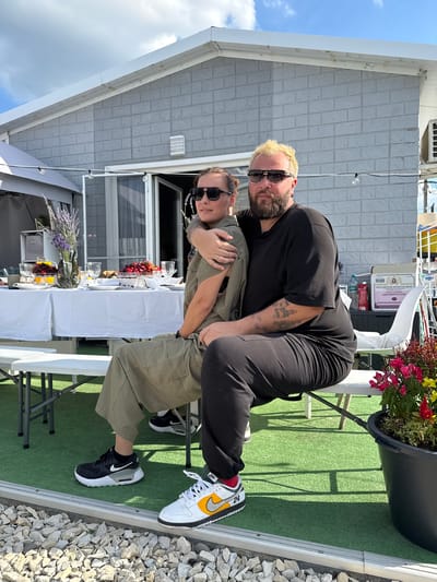 Two individuals are seated closely together on a white bench in an outdoor setting. The person on the left, with short dark hair and wearing sunglasses, is in a reclined position, with their arm around the person on the right. They are wearing a khaki-colored jumpsuit. The person on the right, with blonde hair and sunglasses, has their arm around the other individual. They are wearing a black t-shirt and black pants. Both are wearing athletic shoes.
Behind them, a white-clothed table is set with tableware, glasses, and food, including what appears to be fruit and bread. A vase with purple flowers stands on the table. A gray, brick-patterned building with windows and doors is visible in the background. The ground is covered with green artificial turf and white gravel. The sky is blue with white clouds. The scene appears to be an outdoor dining area. The location is identified as Borisovka, Russia.
