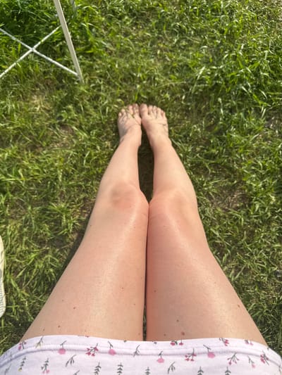 A person's bare legs and feet are visible from a top-down perspective, positioned on a green grassy area. The grass appears to be a mix of short and slightly longer blades, with some patches of dry earth visible. In the upper left corner of the image, a white metal folding chair or support structure is partially visible, its legs crossed. The lighting suggests it is daytime, with the sun casting shadows. The person is wearing light-colored shorts with a pink floral pattern around the hem. The skin on the legs appears slightly tanned and shiny, possibly from sunscreen or sweat. The toes of the feet are spread slightly. The overall scene depicts a moment of relaxation outdoors on a sunny day. The location is not specified in the image or the provided context of Мокино, Russia.