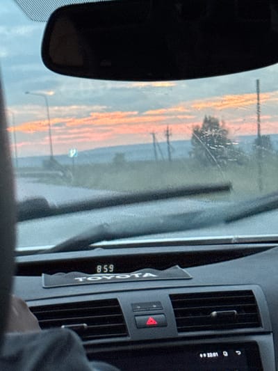 The image shows a view from inside a car looking out the windshield at sunset. The sky is filled with pink and orange clouds. In the distance, there are hills and trees. The car's dashboard is visible in the foreground, with a clock displaying "22:01". The word "TOYOTA" is visible on the dashboard. The scene is consistent with a roadside location in the vicinity of Подымалово, Russia, given the time of day and the rural landscape.