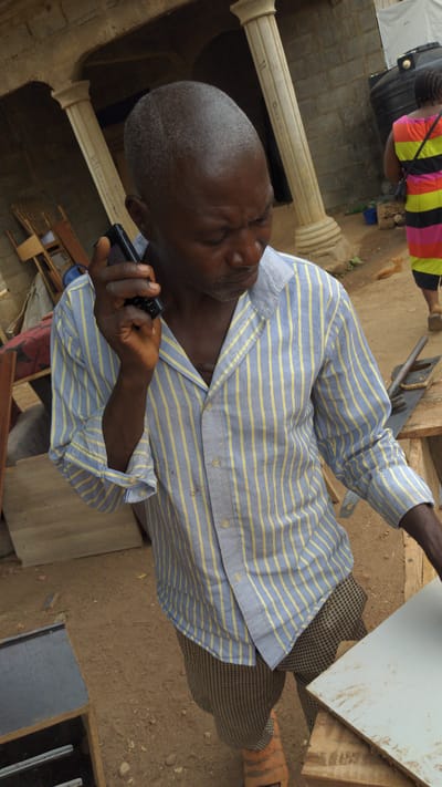 An overhead, rotated photograph captures a man interacting with a feature phone in what appears to be an outdoor or semi-outdoor workshop environment in Masaka, Nigeria.
The main subject is a man with dark skin, closely cropped greying hair, and a slight beard. He is wearing a long-sleeved, blue and yellow striped shirt, partially unbuttoned, and brown/black checkered trousers. He holds a black feature phone with a physical keypad in his right hand, his eyes squinted as if looking at the screen or into sunlight.
In the background, another person, with their back to the camera, wears a vibrant striped shirt (pink, orange, yellow, black) and dark pants, carrying a black bag. Further back, a leg with an orange-brown shoe or sandal is visible.
The setting includes bare, dusty ground and several architectural elements like two light-colored, ornate pillars. There are also various wooden materials, including planks and what appears to be a saw, suggesting a carpentry or construction area. A white rectangular panel, possibly a board, rests on wooden blocks.
The scene appears to be taking place during the daytime, under bright, sunny conditions. On one of the pillars, faint handwritten text is visible, including what looks like "OARAA" and a "Q" or "O".