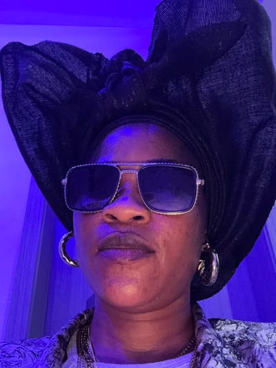 A woman in Ajara, Nigeria, is pictured from the chest up, wearing a black headwrap, oversized square sunglasses with silver trim, and large hoop earrings. Her top is patterned with black and white designs. The lighting is predominantly purple, casting a shadow on the left side of her face. She is looking directly at the camera.