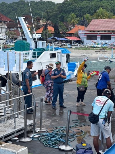 In Manggis, Indonesia, at a ferry terminal, several individuals are gathered on a wet, paved surface. A large white ferry, with the name "SAMY" visible on its superstructure, is docked. The ferry has a blue and white cabin and a white crane arm. In the background, lush green hills rise above a cluster of buildings with red-tiled roofs. Several traditional Indonesian outrigger boats are pulled up on the sandy beach.
Two men in dark blue uniforms, likely port authority or security personnel, stand on the dock. One man, facing left, is speaking, with his right hand slightly raised. The other man, facing right, stands with his hands clasped in front of him.
A woman in a bright yellow jacket over a green shirt, black pants, and sandals is smiling and holding up two bottles in her right hand and two colorful cans in her left hand. She wears a brown crossbody bag.
Another man in a blue shirt and a baseball cap is visible to the right of the woman, facing away from the camera. He is wearing glasses and has a black bag slung over his shoulder.
A third man in a light blue t-shirt with the text "WIJAYA PERKASA Fast Boat" on the back, and white shorts, is also facing away from the camera. He is wearing a black bag and has a water bottle clipped to his belt. He appears to be engaged in an activity, possibly related to cleaning or maintenance, as a black trash bag is near him.
Several stanchions with silver poles and black bases are positioned on the dock, some with red ropes attached, suggesting a queuing area. A coiled blue hose lies on the ground, and a large blue plastic container with a yellow cap is present. The ground is wet, indicating recent rain or damp conditions. The overall scene depicts a busy port environment, likely a departure or arrival point for ferry services.