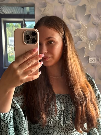A young woman with long, reddish-brown hair is taking a mirror selfie in a room. She is holding a light pink smartphone up to the mirror with her left hand. Her nails are painted with a light pink French manicure. She is wearing a light blue, off-the-shoulder top with a black leopard print pattern. A delicate, thin necklace is visible around her neck. The background of the room includes a window with greenery visible outside, and patterned wallpaper on the wall. The overall scene suggests an indoor, domestic setting. There is no indication of the city of Nizhnyaya Gar', Russia within the image.