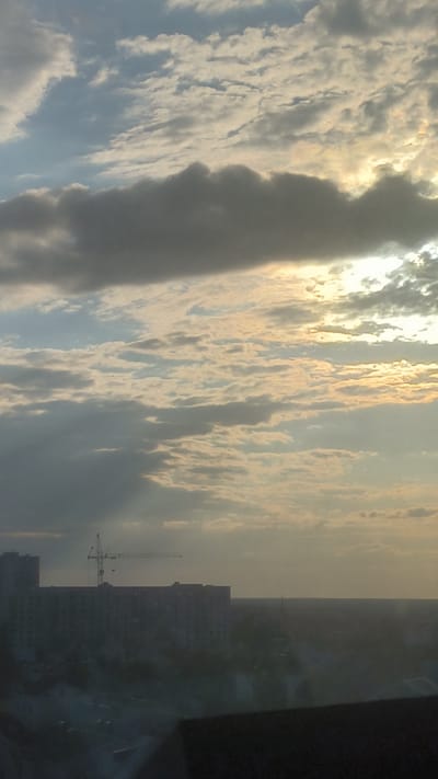 A cloudy sky with scattered sunlight filtering through the clouds is visible in the upper portion of the image. Below the sky, a silhouetted cityscape is depicted. In the middle ground, a long, multi-story building is prominent, with a construction crane situated on top of it. To the left of this building, another, taller building is partially visible. The lower foreground shows dark shapes that appear to be parts of buildings or structures, possibly from a lower vantage point. The overall scene suggests an urban environment, likely under construction or undergoing development, under an overcast sky with shafts of light. The provided crops highlight the cloud formations and the urban structures in more detail.