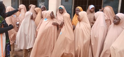 In Unguwar Shanu, Nigeria, a group of approximately ten school-aged girls, all wearing light tan hijabs and modest dresses, are gathered outdoors near a building with a barred window. One adult woman, dressed in black with a colorful patterned scarf, stands to the left and appears to be addressing the girls, pointing with her left hand towards them while holding a black smartphone in her right. The girls are facing the woman, with some looking at her and others looking away. Several of the girls have badges with blue and white emblems on their hijabs. The environment suggests an educational or community setting. The lighting indicates it is daytime.
