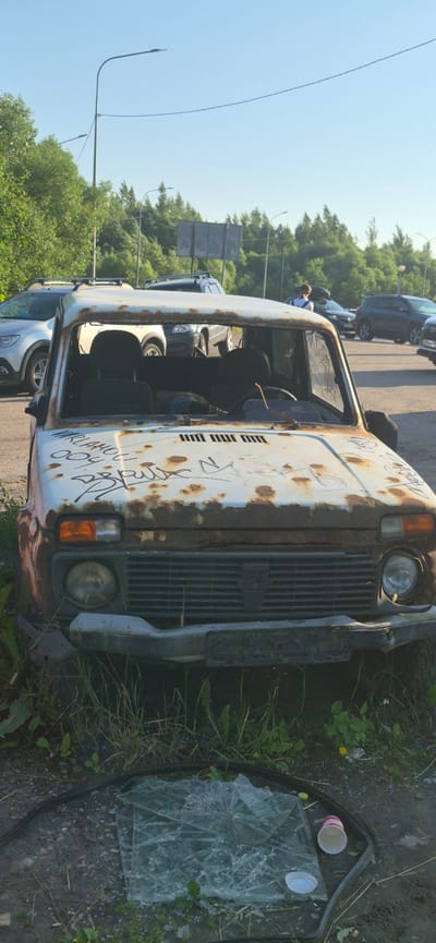 The image depicts a scene in Pushkin, Russia, featuring a heavily rusted, derelict white car in the foreground. The car appears abandoned, with its doors open, revealing dark seats. The exterior is covered in graffiti, including what appears to be Russian text and numbers. The front of the car, including the grille and headlights, is visible. To the right of the car, on the ground, is a pile of shattered glass and a disposable cup. The background shows a road with other cars, some of which appear to be in better condition. Trees and foliage are present, suggesting an outdoor setting. The sky is clear and blue, indicating daytime. The overall impression is one of decay and neglect. Some visible text on the car reads "VARLAMOV 004" and other stylized graffiti.