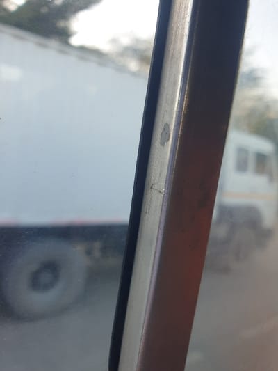 A close-up shot captures a weathered window frame. The frame is a combination of a dark brown upper portion and a silver metal strip along the bottom. There's a thin black rubber or sealant strip between the metal and glass. Scratches and paint chipping are visible, adding to the frame's aged appearance. A blurred reflection of a vehicle is seen through the glass.