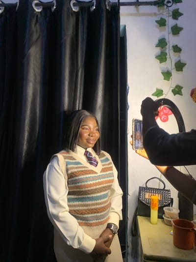 The image shows a young Black woman posing for a photograph in what appears to be an indoor setting. She is wearing a light-colored collared shirt with a patterned sweater vest over it and a striped tie. She has long, dark hair and is smiling at the camera. The background features a black curtain and a white wall adorned with artificial green ivy. In the upper portion of the image, a person's arms are visible holding a smartphone, seemingly taking the picture. A ring light is also present, positioned above the phone. To the right of the phone, there are a few objects on a light-colored surface, including a small red cup, a clear plastic container, and a small, textured bag with a yellow cylindrical object. The lighting suggests it is daytime, but the indoor setting makes it difficult to ascertain specific weather conditions. The overall mood appears cheerful and focused on capturing a portrait. The location is identified as Gwafan, Nigeria.