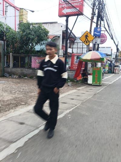 A blurry image shows a person walking on a sidewalk next to a street. The individual wears a dark-colored jacket with lighter-colored sleeves and collar, paired with dark pants and shoes. Behind the person, there's a scene of urban life with power lines, street signs, and a red "SUPERMARKET" sign with an arrow. A yellow pedestrian crossing sign is visible along with a snack stand covered by an umbrella. The background includes buildings and trees.