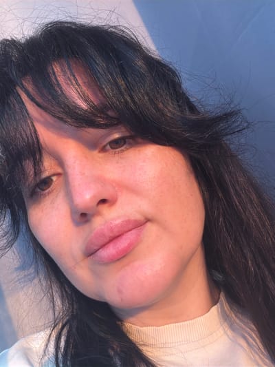 The image is a close-up, high-angle selfie of a young woman with dark hair and light skin, exhibiting a fair amount of freckles, particularly on her cheeks and nose. Her eyes are a warm brown, and her lips are full and slightly parted, appearing to have a subtle pink tint, possibly from lip balm or natural color. Her expression is relaxed, with a slight tilt of her head.

The lighting in the image is warm and directional, casting a soft glow on her face and highlighting the texture of her skin. This lighting creates subtle shadows and emphasizes the contours of her face. Her dark hair is slightly disheveled, with some strands falling across her forehead and face.

She is wearing a light-colored, possibly cream or off-white, ribbed crewneck shirt. The background is a gradient of soft blue and pink hues, with diagonal lines of light and shadow suggesting a window or sheer curtain.

There is no identifiable information within the image that would indicate a specific location in the city of Korolyov, Russia. The setting appears to be indoors or in a sheltered outdoor space with a diffused light source.