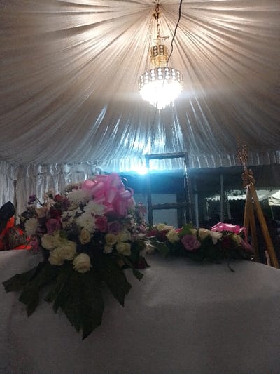 The image displays the interior of a draped tent structure within the city of Marenyo-Shianda ward, Kenya. A multi-tiered, gold-colored chandelier with crystalline elements hangs centrally from the pleated white fabric ceiling, emitting bright light. In the foreground, a substantial floral arrangement featuring white, pink, and mauve roses, along with other white filler flowers and green foliage, is positioned on a white, fabric-covered surface. A large pink bow is prominently integrated into the floral display. To the left, a dark, indistinct object is partially visible. In the middle ground, a dark, skeletal frame or structure is visible. A bright blue-white light source is present in the background, emanating from beyond the immediate tent interior. To the far right, an ornate golden object on a stand is partially visible. No individuals are clearly identifiable.