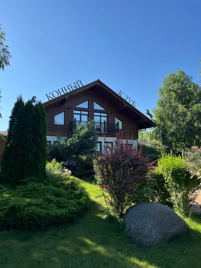 This is a full, eye-level outdoor shot of a wooden building with a white lower level. The building has a pitched roof with a sign on it that reads "КОННЫЙ КЛУБ" in metal letters, and a metal horse statue on the peak of the roof. There are trees and shrubs in front of the building, and a large grey boulder is in the foreground. The sky is clear and blue, and the sun is shining, casting shadows on the grass. The image appears to be taken during the day. There are no people visible in the image. The building is situated in a verdant, landscaped setting, indicative of a recreational facility. The location is Plastunivska, Russia.
