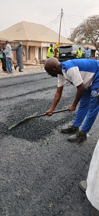 The image captures an outdoor scene depicting road construction or repair activities under bright daylight.

In the foreground, a man with a bald head, wearing a white t-shirt with blue sleeves and collar, and blue track pants with a white logo on the left thigh, is bent over. He is actively using a long, slender stick or tool, held with both hands, to spread or level a dark, granular material, likely asphalt or gravel, on the ground. His dark shoes are visible as he stands on the material.

The ground where the activity is taking place is covered with this dark, rough-textured material, indicating a newly laid or repaired surface. Further into the background on the right side, the dark material extends, suggesting a road or pathway being constructed. The feet of another person, wearing dark shoes and light-colored attire, are partially visible in the upper right, standing on this surface.

On the left side of the image, several other individuals are present. Some are visible closer to the background, wearing high-visibility yellow or green vests and possibly hard hats, suggesting they are part of the construction crew or supervisors. Behind them, a dark-colored vehicle, possibly an SUV or pickup truck, is parked. Further left, there is a light-colored, single-story building with a light roof, appearing somewhat weathered. A wooden utility pole with multiple wires extending from it stands in front of this building. Closer to the bottom left, other people are visible, dressed in a mix of traditional long robes and casual wear like a plaid shirt and jeans. A tree with bare branches is discernible behind the building, hinting at a dry season or a specific type of deciduous tree.

The overall scene suggests a community effort or professional roadwork being carried out in what appears to be a populated area, given the presence of buildings and multiple individuals. The weather appears clear and dry, with no strong shadows, possibly indicating an overcast sky or hazy conditions. No explicit text is clearly visible other than a potential logo on the main subject's pants.