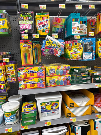 The image captures a brightly lit aisle within a retail store, heavily stocked with various art and craft supplies. No people are visible, and the scene implies a typical shopping environment.

The shelves and pegboards are filled with an array of products, including numerous markers such as Crayola Color Change, Crayola Bold & Bright Washable Markers, Funky Chunky Mini Markers, Mr. Sketch Scented markers, and Crayola Silly Scents Smashups. Colored pencils are also prominent, with boxes of sharpened pencils from CraZArt and Scholar (Manga pencils) displayed.

Paint supplies include multiple boxes of CraZArt Washable Project Paint (Classic), Crayola Washable Paint, and sets of CraZArt Artist Brushes. Modeling materials like large tubs of Crayola Model Magic and Crayola Air-Dry Clay are positioned on lower shelves. Various pen+GEAR Sketch Pads and Sketch Books are stacked neatly in the upper right section of the image.

Price tags are visible throughout the display, indicating costs for individual items, such as $5.97, $4.97, $9.67, $2.44, $4.57, $20.93, $5.88, $3.96, and $10.62. The organization and variety suggest a dedicated section for children's art supplies or general creative materials. The lighting is uniform, characteristic of an indoor retail setting.