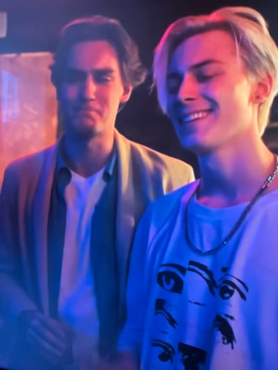 The image shows two young men in an indoor setting, possibly a club or a party, bathed in blue and pink neon lights. On the left, a man with dark hair, wearing a beige blazer over a blue shirt and a white t-shirt, looks towards the camera with a neutral expression. On the right, a man with blond hair, wearing a white t-shirt with a graphic print of eyes and a silver chain necklace, smiles broadly with his eyes closed. The background is dark and out of focus, with some blurry elements that suggest a curtain or fabric. There is no discernible context linking this image to Vangaži, Latvia.