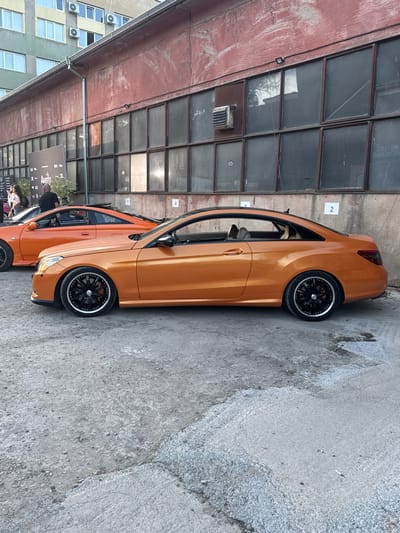 Two orange sports cars are parked on a cracked asphalt surface in front of a red industrial building with large, grimy windows. The building has a weathered facade. In the background, a taller, multi-story building with visible air conditioning units is visible. The scene appears to be an outdoor event or gathering, possibly a car show, given the presence of a banner with logos and text on the left side of the image and a number "2" affixed to the wall of the industrial building. The lighting suggests it is daytime, with some sunlight reflecting off the windows. The ground is littered with small debris. No people are clearly identifiable or engaged in any specific actions in the main focus of the image. The context provided about Busmantsi, Bulgaria, indicates a potential industrial or outskirts area of the city.