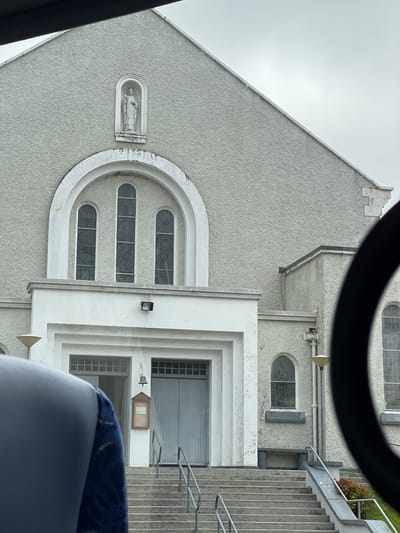 The image shows the facade of a church in Gortahork, Ireland. The church is constructed of light gray stucco and features a large arched window with three stained-glass panes above the main entrance. A statue of a woman in a niche is positioned above the arched window. The entrance is framed by a white portico, and a set of concrete stairs with metal railings leads up to double doors. To the right of the main entrance, a smaller arched window is visible, as well as a lamppost. Part of a dark, curved object, possibly a vehicle's interior, is in the foreground, obscuring the lower left portion of the image and a portion of the right side. The sky is overcast.