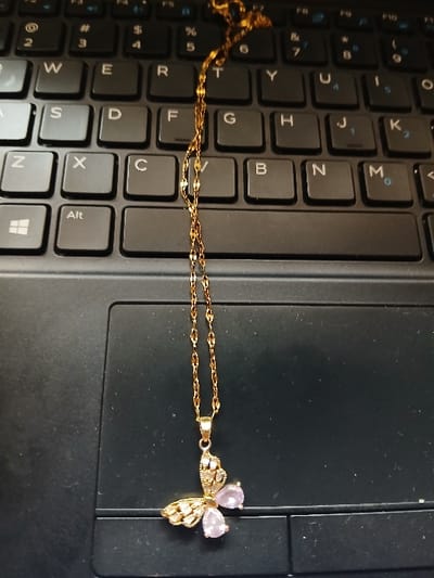 A gold-colored necklace with a butterfly pendant rests on a black laptop keyboard and touchpad. The delicate chain is draped across the upper keys of the keyboard. The butterfly pendant features one wing encrusted with small, clear stones and the opposing wing adorned with three teardrop-shaped, light purple stones. The pendant itself lies on the lower-left area, partially covering the 'Alt' key and extending onto the dark grey touchpad surface. Visible keyboard keys include 'Q', 'W', 'E', 'R', 'T', 'Y', 'U', 'I', 'O', 'P', 'A', 'S', 'D', 'F', 'G', 'H', 'J', 'K', 'L', 'Z', 'X', 'C', 'V', 'B', 'N', 'M', along with numeric and symbolic keys, and a Windows logo key. No individuals are present in the scene. The environment is consistent with an indoor setting. The specific location within Witeithie ward, Kenya, is not visually verifiable from the image.