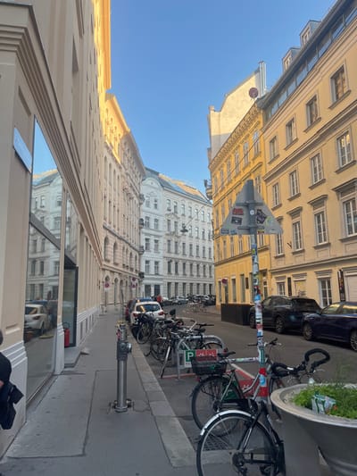 The image shows a street in Vienna, Austria, located at 48.202 latitude and 16.351 longitude. The street is lined with tall buildings with yellow facades and is relatively empty, with only a few people present. There are several bicycles parked on the sidewalk next to a tall, metallic fire hydrant. The buildings on the right side of the street are yellow, while the buildings on the left are white. There is a tall building with a large, rectangular glass window that is reflecting the surrounding street. The street is paved with gray cobblestones, and the buildings are all multi-story with balconies, windows, and doors. The weather appears to be sunny and clear.  The image is taken from the sidewalk, looking down the street.