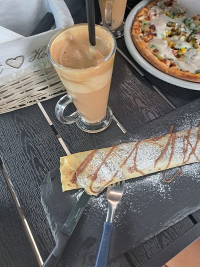 A close-up, overhead shot shows a table setting with a pizza and a crepe. The pizza, partially visible in the upper left, is a round, thick-crust variety topped with melted cheese, corn, peas, and possibly pickled cucumbers and meat. Next to the pizza is a tall, clear glass filled with a light brown, frothy beverage, likely a milkshake or coffee drink, with a black straw. To the right of the drink, on a dark slate board, lies a crepe dusted with powdered sugar and drizzled with chocolate sauce. A fork and a small knife, with "TRAMONTINA MADE IN BRAZIL" printed on it, rest on the slate board next to the crepe. The table appears to be dark wood with a textured surface. A white woven basket and a white napkin with embroidered hearts and script lettering are visible in the lower left. The scene suggests a casual meal or snack, possibly outdoors given the natural lighting. The overall impression is one of a leisurely moment.