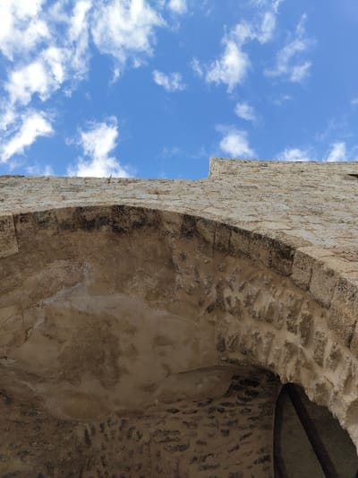 A weathered stone archway curves across the frame, its rough, aged texture contrasting with a bright blue sky dotted with fluffy white clouds. The stone is a light, creamy color with darker, shadowed areas highlighting the individual blocks. A dark horizontal beam sits just inside the upper arch. The structure appears old and possibly part of a larger building. The perspective is from below, looking upwards towards the sky.
